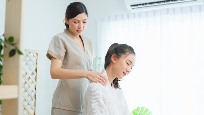 亚洲年轻快乐女性在肩部和背部按摩时感到放松。美丽迷人的女孩坐在按摩台上，在spa美容院接受按摩师的理