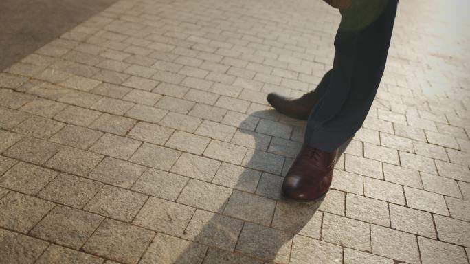 视频中，一位商人的脚穿着棕色皮鞋，在铺好的街道上左右移动，而相机在四处飞舞