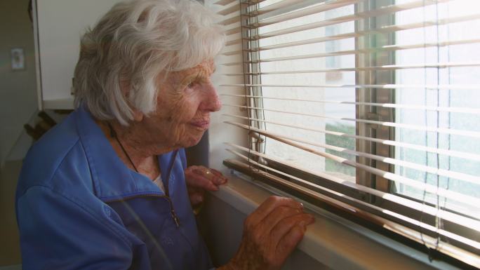 一位年长的白人妇女站在室内透过百叶窗向外看