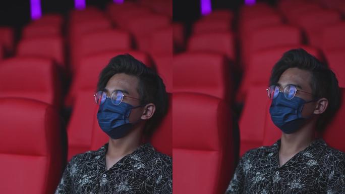 戴着面具的亚裔中国少年独自在电影院看电影，表情呆滞，两腿交叉
