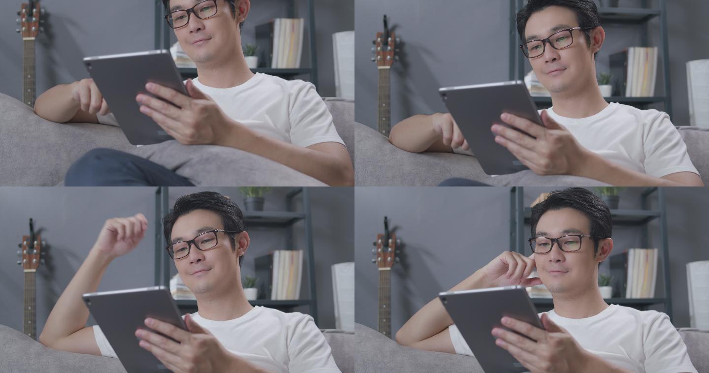 一名亚洲男子坐在家客厅的沙发上，一边在数字平板电脑上阅读新闻。戴眼镜的成年男子在冠状病毒隔离封锁后，