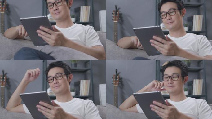 一名亚洲男子坐在家客厅的沙发上，一边在数字平板电脑上阅读新闻。戴眼镜的成年男子在冠状病毒隔离封锁后，