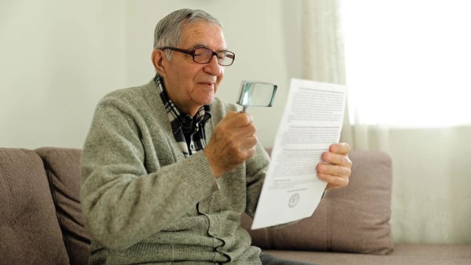 英俊的退休老人戴着灰色开襟羊毛衫眼镜坐在沙发上看报纸，手里拿着loupe室内公寓的照片
