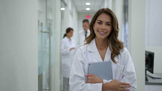 拉丁美洲女医生在医院工作，拿着数码平板电脑微笑着看着摄像机