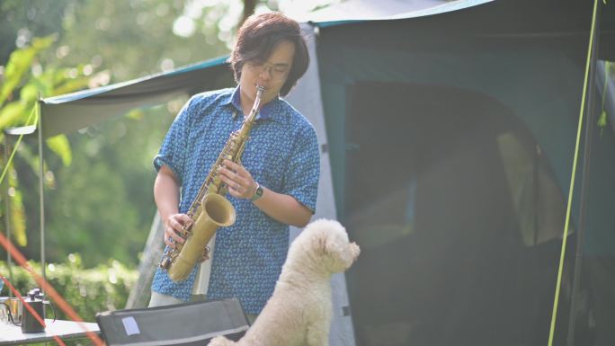 早上，亚裔中国萨克斯管演奏家和他的宠物玩具贵宾犬在露营帐篷前演奏