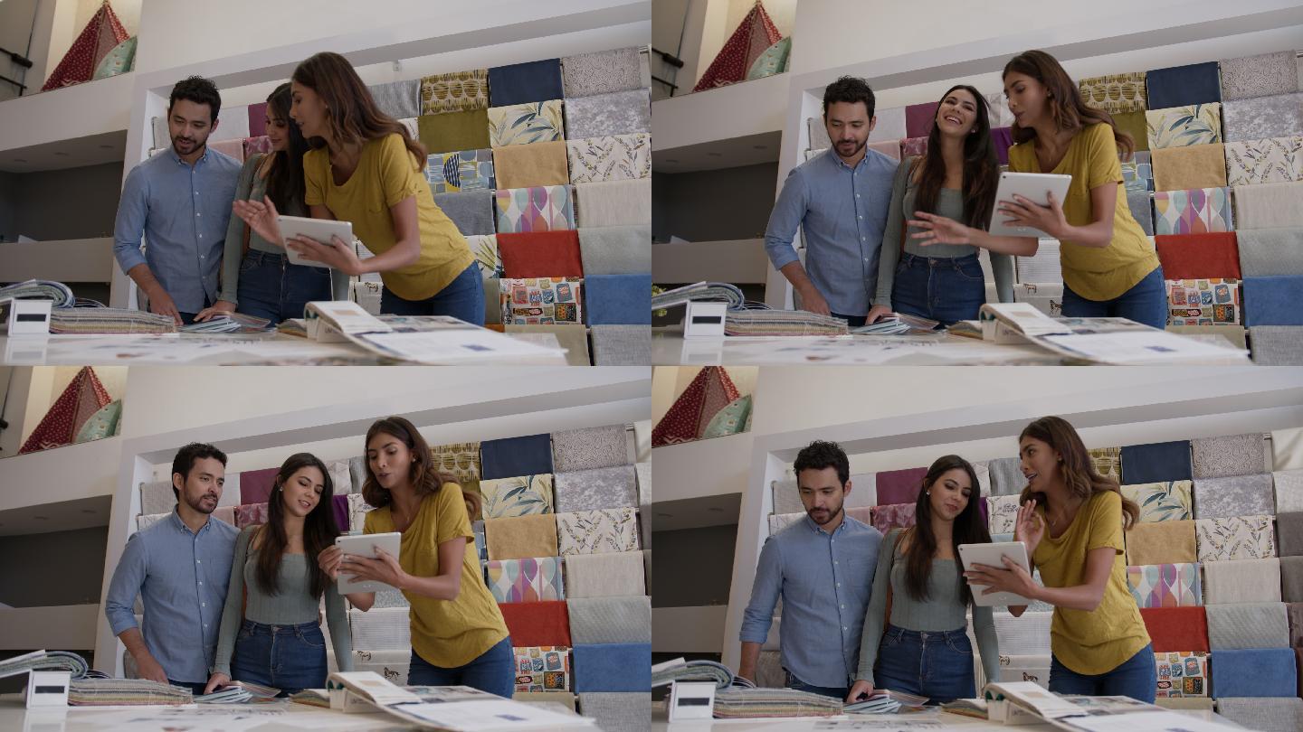 一对幸福的夫妇在友好的女售货员的帮助下为他们的家挑选面料，并向他们展示面料样本和平板电脑上的设计