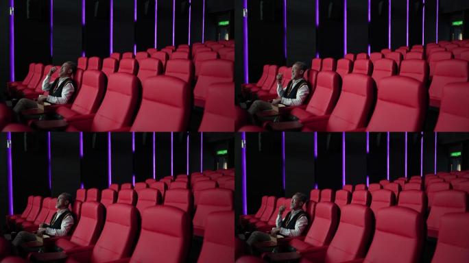 一名留着白胡子的亚裔中国男子坐在空荡荡的电影院红色座位上，一边吃一边看电影。