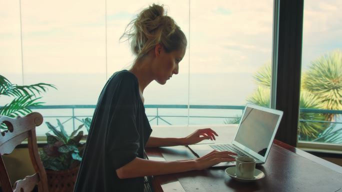在全景窗旁工作的妇女。使用笔记本电脑欣赏史诗般的海景