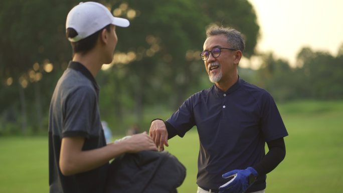 亚洲华人父子高尔夫球手日落时在高尔夫球场散步、谈笑风生