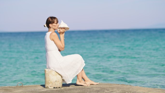 坐在海滩上的一个码头上，斯洛莫年轻女子对着海螺壳吹气