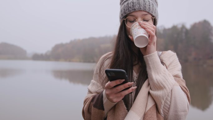 年轻女子在湖边用纸杯喝咖啡时使用手机