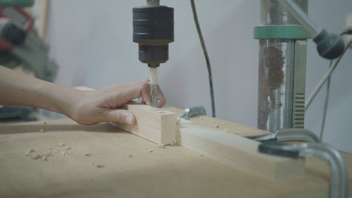一个穿着牛仔裤和格子法兰绒衬衫的女木匠在她的木工车间里用便携式电钻在一块木头上钻孔