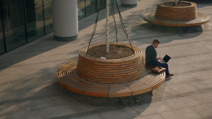 商业中心附近广场的高角度视图，有一个圆形长凳，一个商人拿着笔记本电脑坐在上面