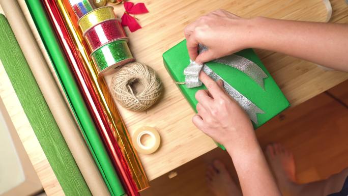 亚洲女性的手用透明胶带将银丝带系在绿色礼品盒上，圣诞礼物放在家里的桌子上，晚上在装饰有圣诞树和装饰品