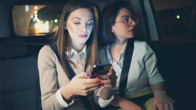 两位兴高采烈的年轻女性坐在汽车后座上使用智能手机