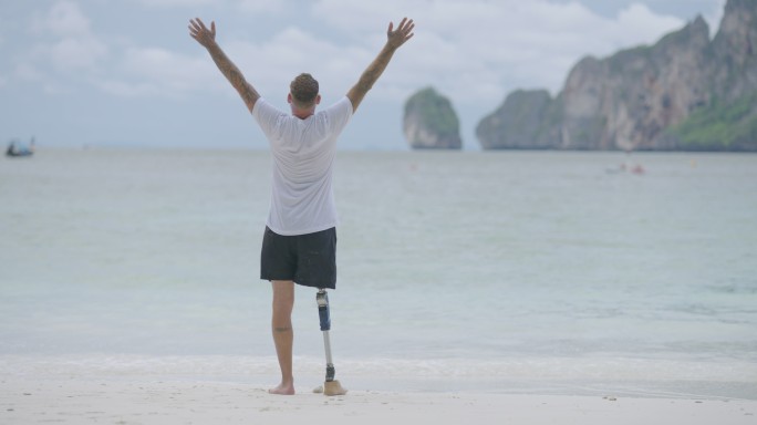 一位残疾人的后视图，他带着假腿，在早上的海滩上张开双臂，向空中举起手臂。残疾收集：日常生活概念。