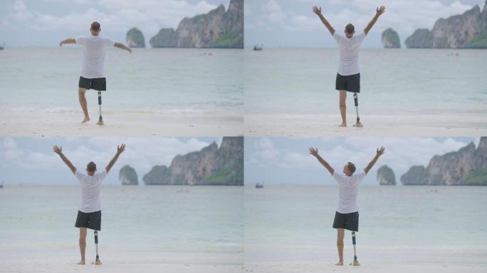 一位残疾人的后视图，他带着假腿，在早上的海滩上张开双臂，向空中举起手臂。残疾收集：日常生活概念。