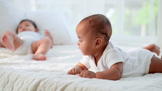 关注一个穿着白色婴儿服的无辜黑人新生儿躺在床上试图爬行，而5个月大的白人新生儿正在踢腿、玩耍，并向后