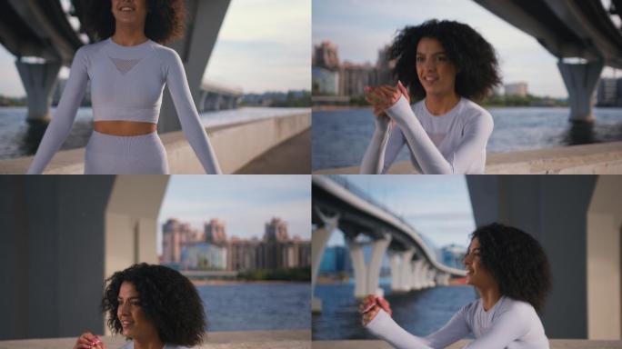 一位身着白色套装的快乐苗条女运动员蹲在现代混凝土桥下的路堤上