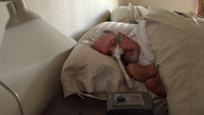 患有睡眠呼吸暂停症的中年男子躺在床上睡觉，他戴着CPAP（持续气道正压通气）机器帮助自己入睡
