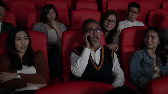 电影在电影院放映时，亚裔中国老人正在使用智能手机。打扰她周围的其他观众