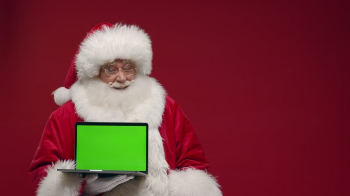 一个快乐的圣诞老人站在红色背景下，微笑着向上竖起大拇指，在相机中展示一个空的笔记本电脑绿色屏幕