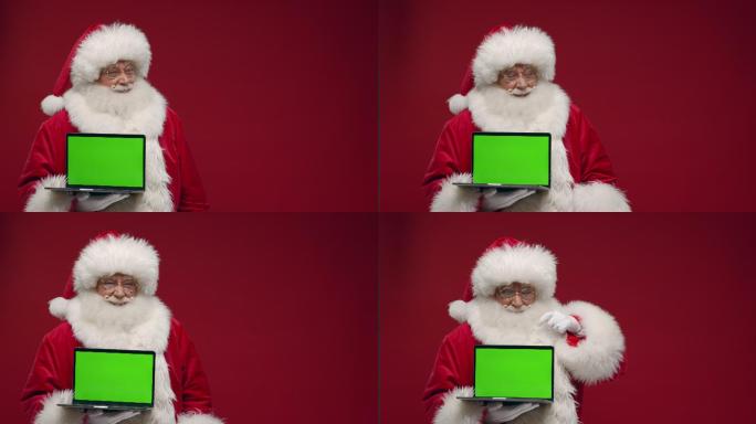 一个快乐的圣诞老人站在红色背景下，微笑着向上竖起大拇指，在相机中展示一个空的笔记本电脑绿色屏幕