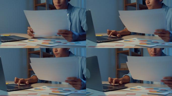 一个亚洲年轻人晚上坐在客厅的办公桌上，拿着笔记本电脑工作的特写镜头。