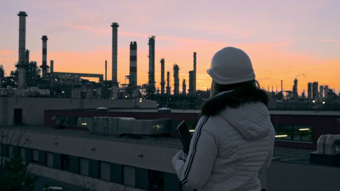 中景：黄昏时分，戴着安全帽的女工程师在炼油厂屋顶上佩戴数字平板电脑