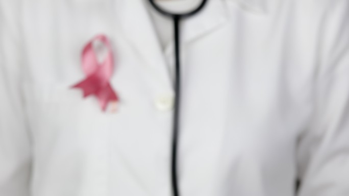 医生戴着粉色乳腺癌意识丝带，将听诊器伸向摄像机进行诊断