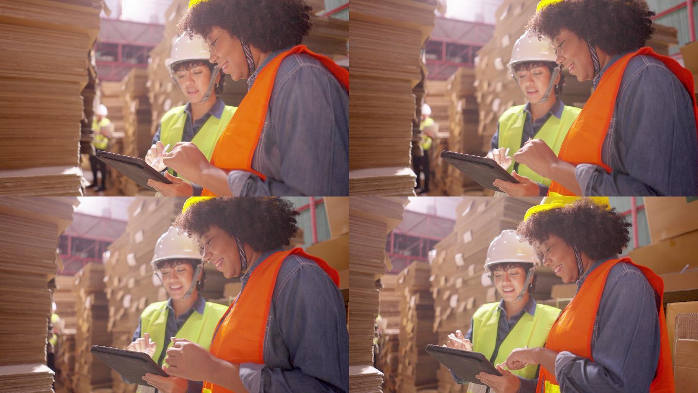 侧视图：在纸箱厂，穿着安全背心和头盔的拉丁和非洲女工们开心地微笑着，用数字平板电脑和智能手机帮助一起