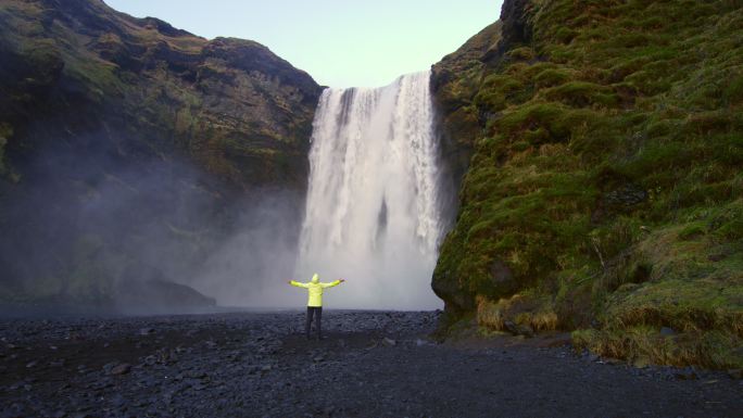 中景：冰岛斯科加福斯瀑布下，一名无忧无虑的女子伸开双臂站在那里