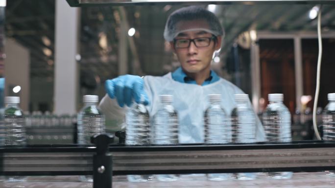 中国亚裔男性生产线工人，穿着干净的劳保用品，在瓶装厂矿泉水饮用水厂检查水瓶