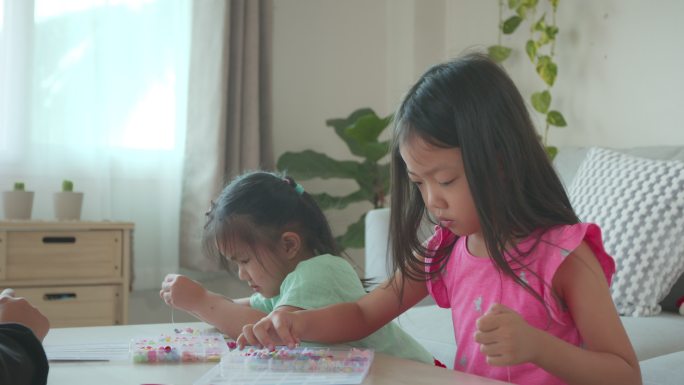 亚洲小女孩和朋友一起在家里把珠子串在绳子上，既有目的又有乐趣。孩子们制作手工手镯来发展手的运动能力