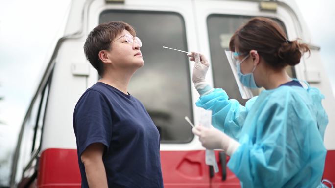 户外救护车旁的亚裔中国女医生带着PPE从患者身上取鼻拭子进行冠状病毒检测。防护套间的医务人员正在用拭