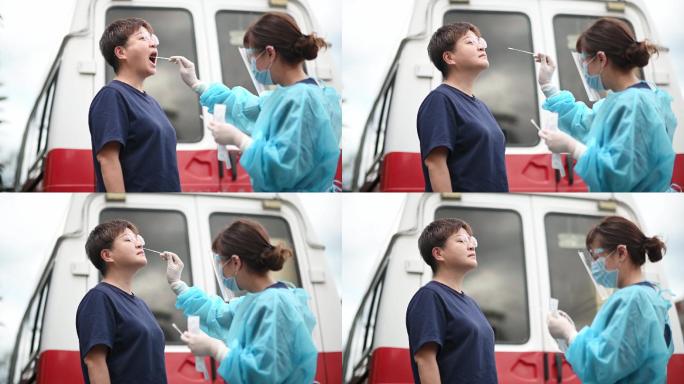 户外救护车旁的亚裔中国女医生带着PPE从患者身上取鼻拭子进行冠状病毒检测。防护套间的医务人员正在用拭