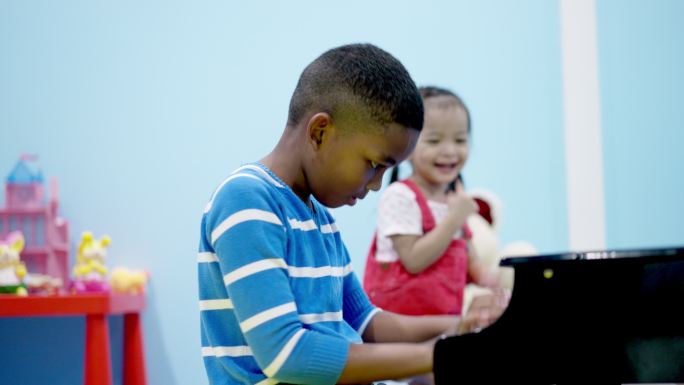 音乐时间，一个非洲黑人孩子，一个7岁的男孩在弹钢琴，而年轻漂亮的亚洲女孩则在微笑，鼓掌，快乐地跳舞。