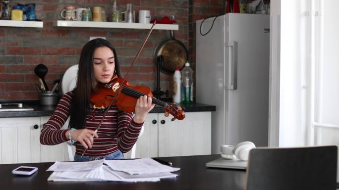 一位年轻的女小提琴手正坐在厨房的桌子旁，看着音符，拉着小提琴。