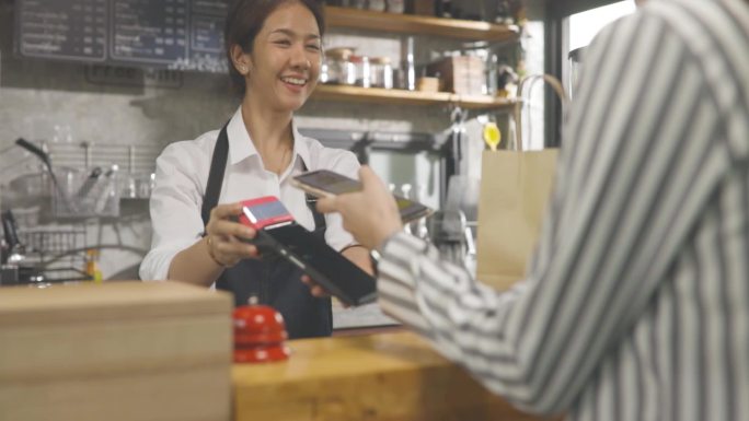 在咖啡馆里，一位美丽的亚洲女士为一位通过手机系统付款的顾客做外卖咖啡