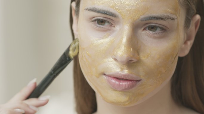 一个年轻的黑发女孩用刷子在皮肤上涂上金色的化妆品面膜。