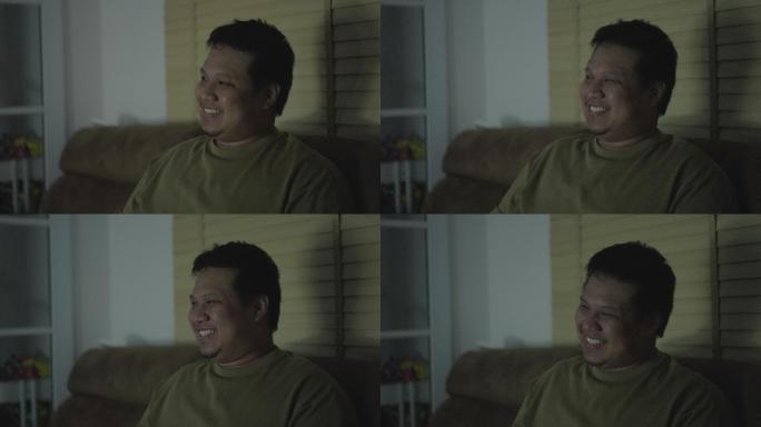 一名亚洲男子晚上在客厅看电视时笑着盯着自己的脸