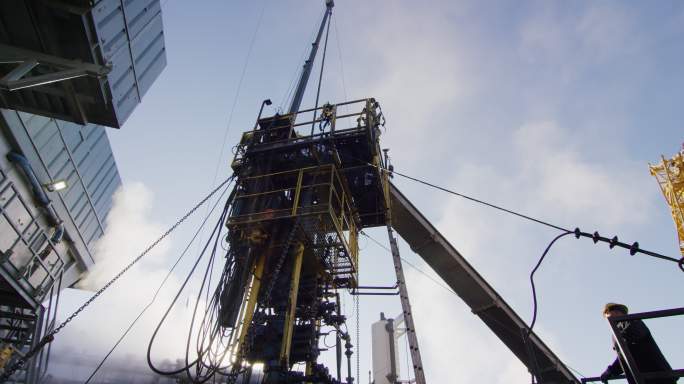 一个寒冷、阳光明媚的冬日早晨，油田工人在油气井场的一个高大的金属平台上装配钻杆的慢镜头