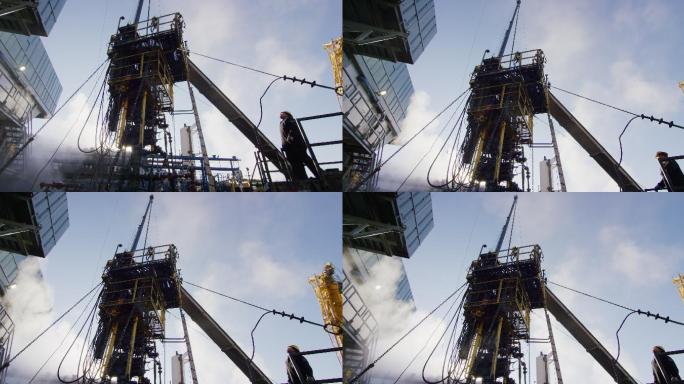 一个寒冷、阳光明媚的冬日早晨，油田工人在油气井场的一个高大的金属平台上装配钻杆的慢镜头
