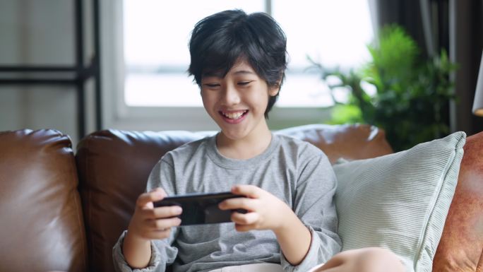 在家客厅的沙发上玩手机游戏的亚洲少年男孩面带微笑，兴高采烈，坐在沙发上玩手机游戏的亚洲少年男性正在玩