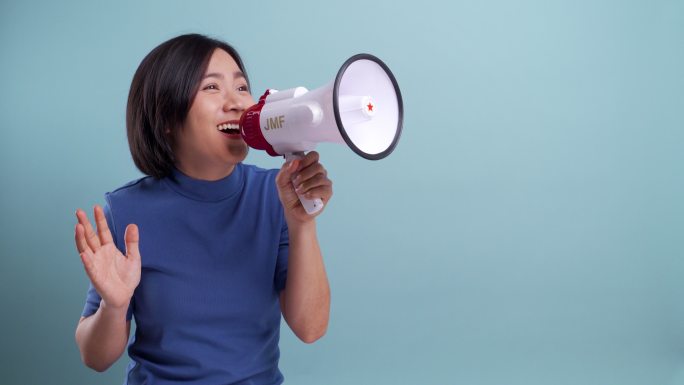 快乐的亚洲女人用扩音器在蓝色背景上做出孤立的叫喊手势。4K视频