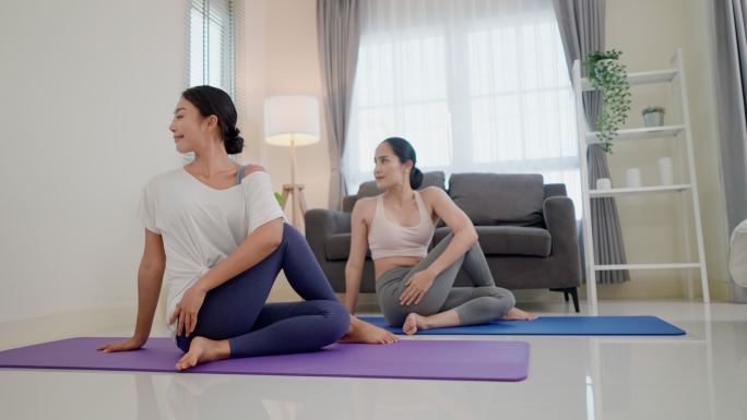 两位亚洲女性做半鱼王瑜伽姿势，右膝弯曲，右脚蜷缩在左臀附近，右臂在右耳旁笔直向上，然后将躯干向左扭转