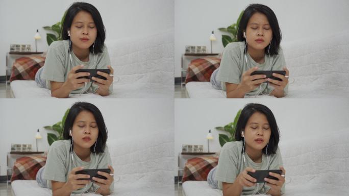 亚洲女性躺着戴耳机在智能手机上玩游戏