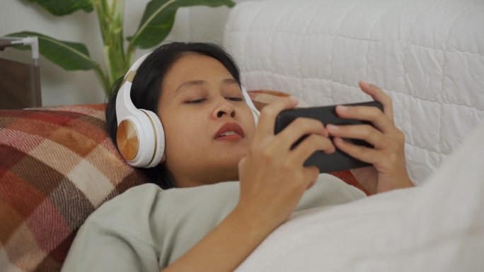 亚洲女性躺着戴无线耳机在智能手机上玩游戏