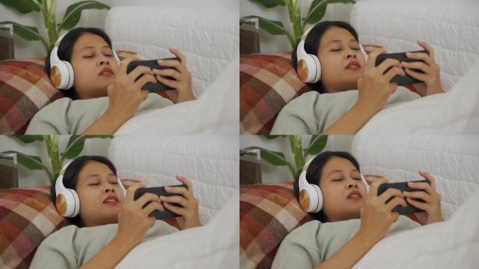 亚洲女性躺着戴无线耳机在智能手机上玩游戏