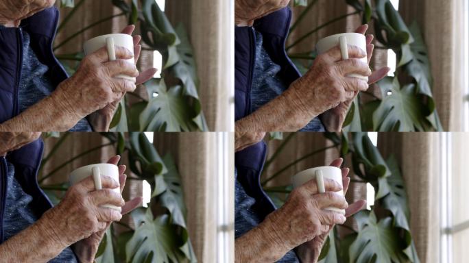 特写镜头：一位年长的高加索人妇女的手拿着咖啡或茶杯，在夏天的室内看着窗外